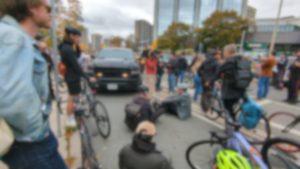 Cyclists sit in the bike lane in front of a large black pickup truck while others stand on the sidewalk. Some are being interviewed by nearby reporters. Image blurred for privacy.