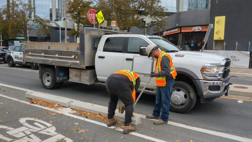 Two construction workers pry out the bolts holding down concrete parking curbs delineating bike lanes. The work truck behind them has the company name and logo covered up.