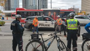 Construction workers with tools in their hands are surrounded by police officers and a cruiser, as a TTC bus passes in the background.