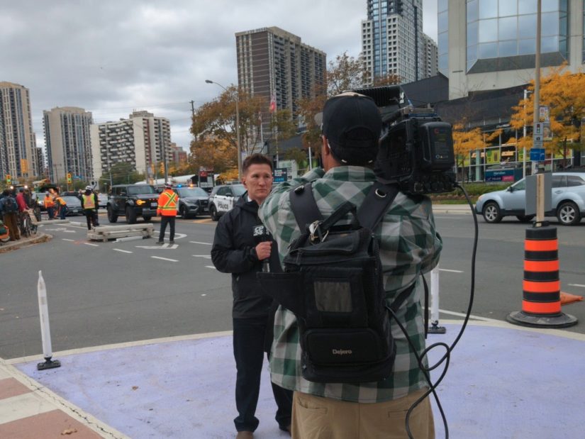 A clean-cut TV reporter speaks into a large microphone while being filmed by a cameraman. The bike lanes are being removed in the background.