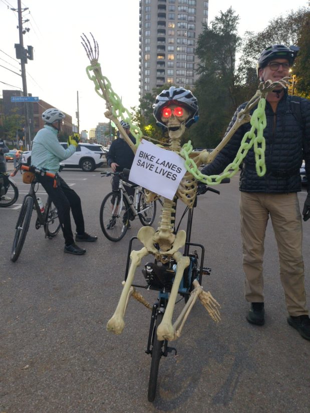 A plastic skeleton Halloween decoration mounted on the front of someone's bike, with glow-in-the-dark chains, a bike helmet, red lights for eyes, and a sign reading BIKE LANES SAVE LIVES.