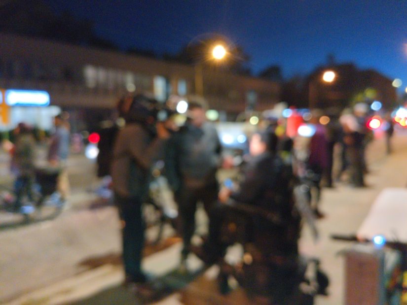 A TV reporter and camera person interview someone in a powered wheelchair. Blurred for privacy.