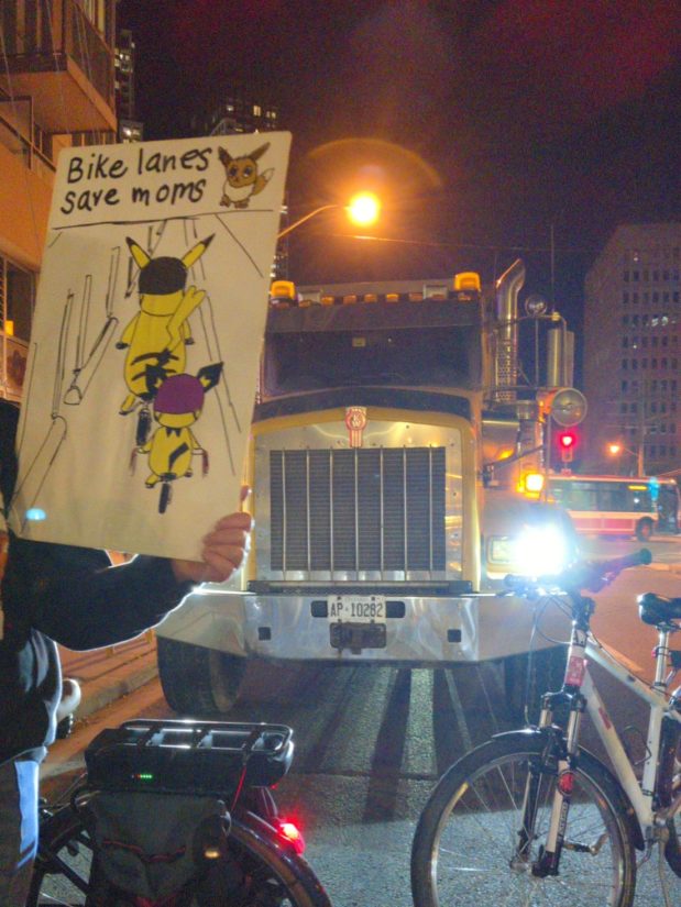 In front of a large truck parked with its lights on, a protester holds up a sign illustrated with various Pokémon saying "Bike lanes save moms".