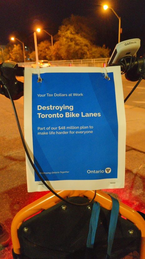 On the front of a cargo bike, a poster made to look like official Ontario ones, reading "Your Tax Dollars At Work Destroying Toronto Bike Lanes Part of our $48 million plan to make life harder for everyone" And in small print: "Destroying Ontario Together" and the Ontario trillium logo.