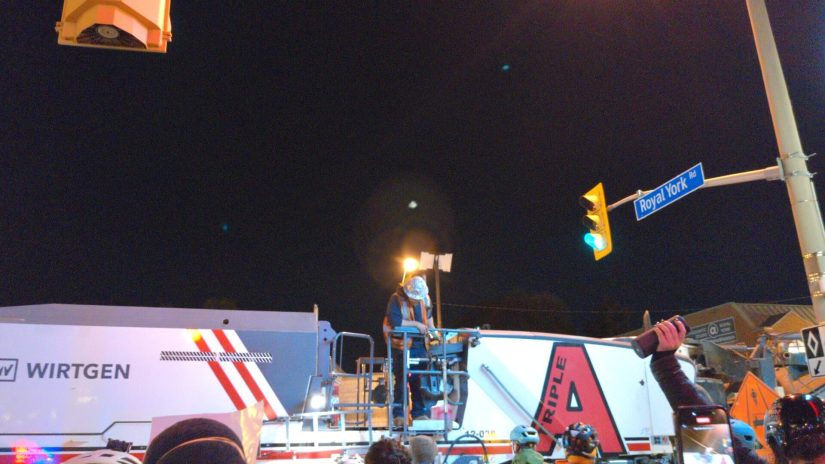 A worker on top of a milling machine looks down at a crowd of protesters, one of whom is holding up a Bluetooth speaker.