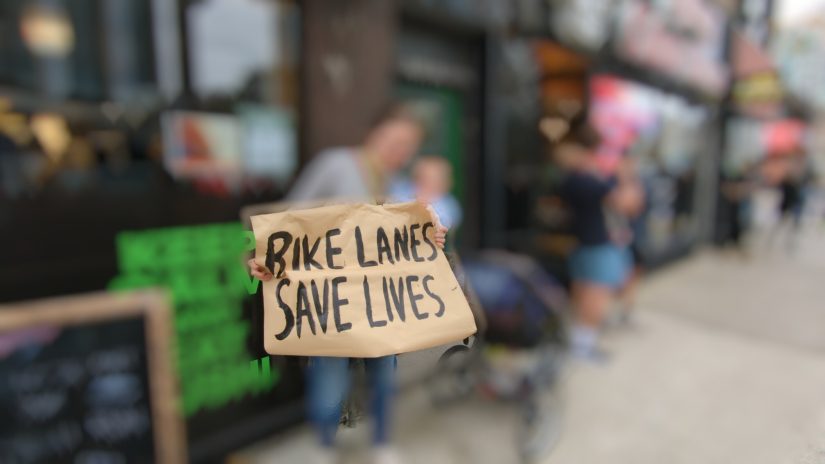 A parent with a baby on their hip holds up a hand-painted sign on brown paper reading BIKE LANES SAVE LIVES. All but the sign blurred for privacy.