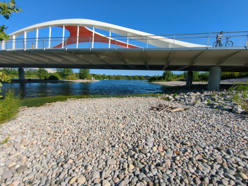 A swooping white and red bridge passes over the Don, with a beach of rounded, fist-size rocks in the foreground.